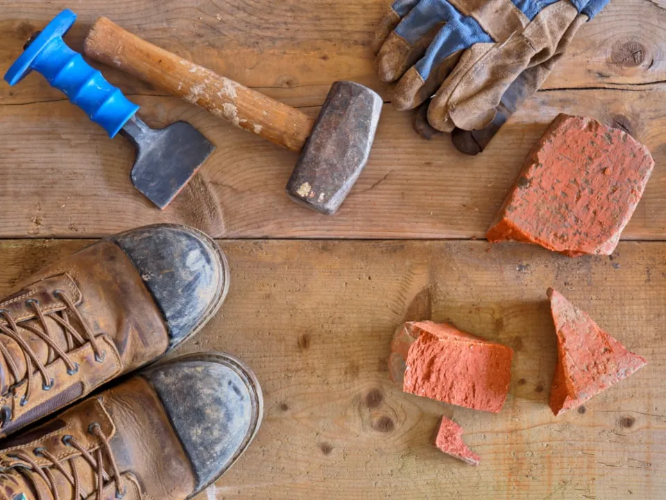 Work boots, gloves, a club hammer and a bolster beside broken bricks on timber boards