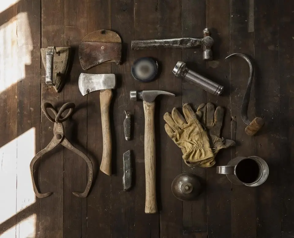 Old hand tools laid out on dark timber boards: a hammer, a hatchet, tongs, a tape and a pair of leather gloves
